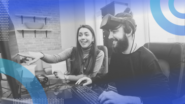 Play framework image of two game developers working at a computer together. The white woman on the left has long dark hair and wears a casual long sleeve t-shirt. She smiles and points to the screen. The white man on the right wears a long-sleeved black shirt with the sleeves pushed up. He wears headphones and a 3D gaming device on the top of his head. He is also smiling.
