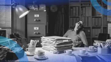 A woman sleeps at a desk piled with paperwork, coffee cups, and other detritus