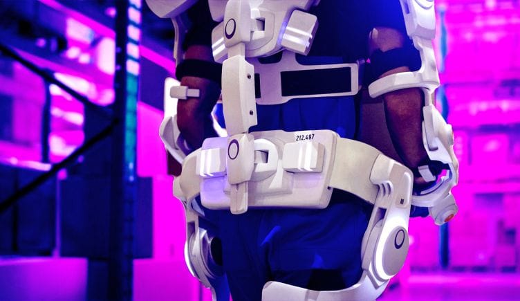 A person carrying boxes with the support of an exoskeleton on their back and limbs.