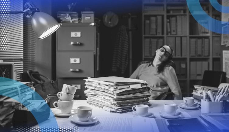 A woman sleeps at a desk piled with paperwork, coffee cups, and other detritus