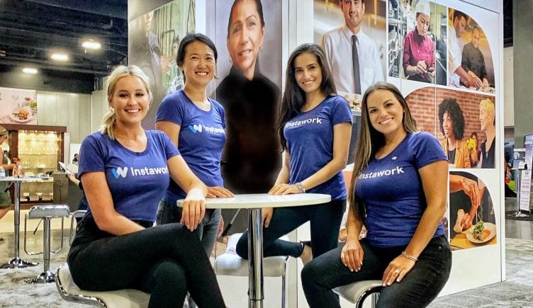 Instawork employees in branded apparel sit in front of a conference display.