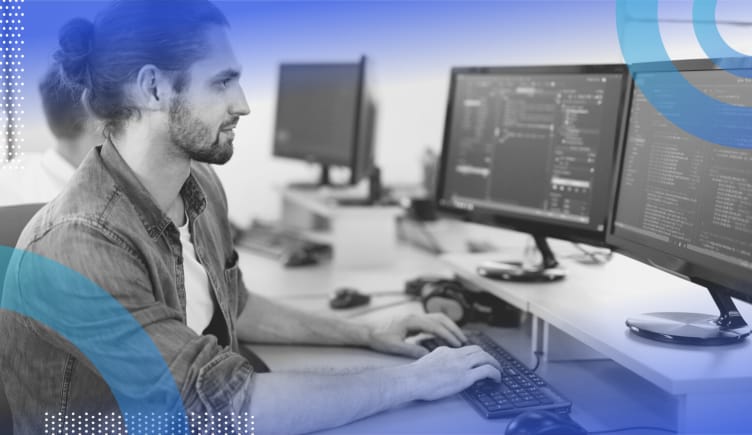 Ember.js image of a white man with a trimmed beard and his hair pulled back in a bun. He types code on a dual-monitor system. He wears a chambray shirt.