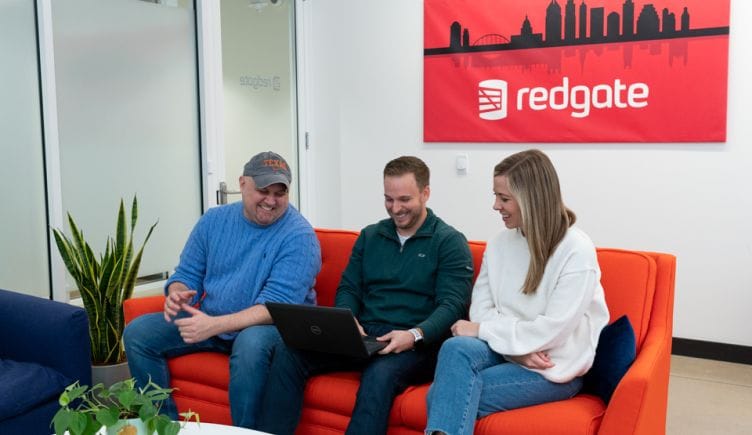 Three Redgate employees sitting on a red couch with a Redgate picture and logo behind them.