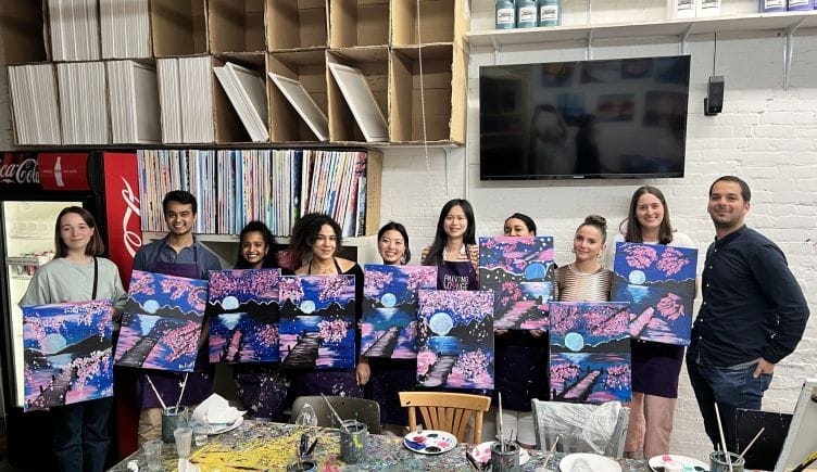 Some of Torch's employees posing for a photo, each holding their own painting of a body of water reflecting the moon and surrounded by cherry blossoms.