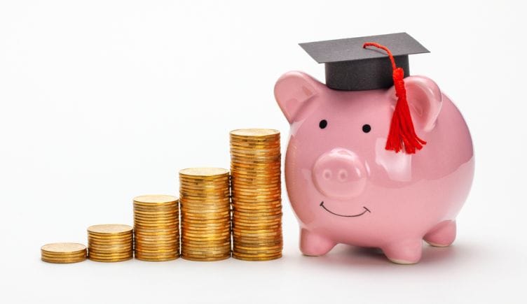 A graduation cap sits on top of piggy bank next to stacks of coins.