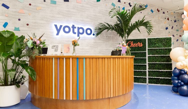 Photo of Yotpo’s front desk surrounded by plants with lighted logo sign on the wall behind