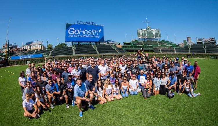 Large group photo of GoHealth team in Wrigley Field’s outfield with GoHealth logo on billboard behind them.