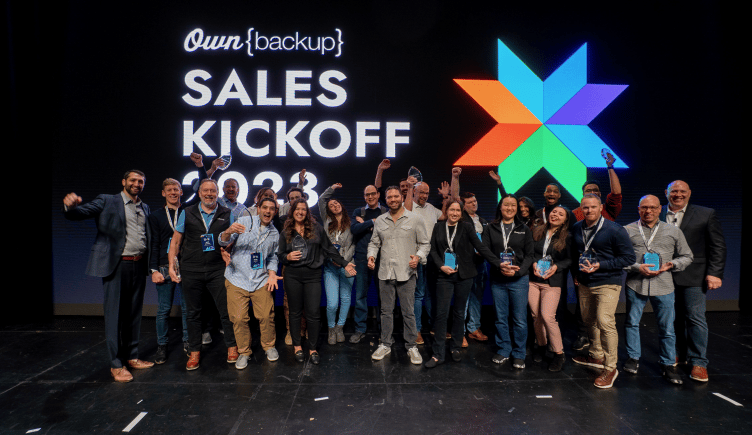 Photo of OwnBackup team members on a stage with large screen behind reading “OwnBackup Sales Kickoff 2023.”