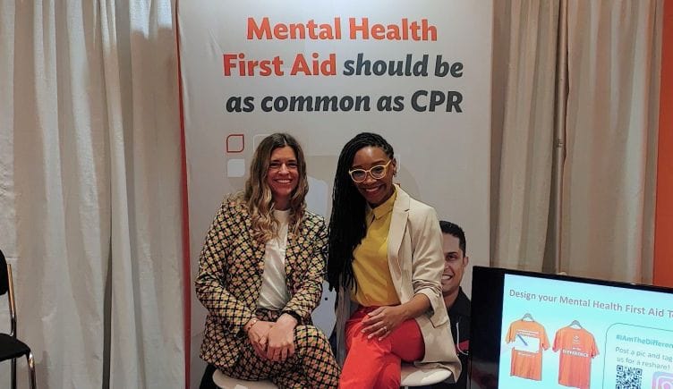 Photo of two Tapestry team members at a booth in front of banner that reads “Mental Health First Aid should be as common as CPR”
