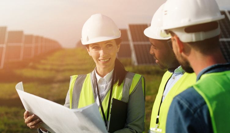 Three engineers wearing hard hats and work vests stand together looking over blueprints in a field surrounded by solar panels.