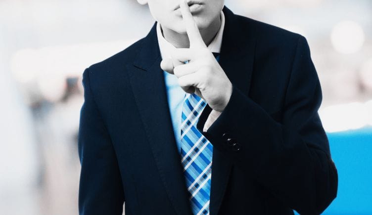 A person in a suit puts a finger to their lips as if to say, 