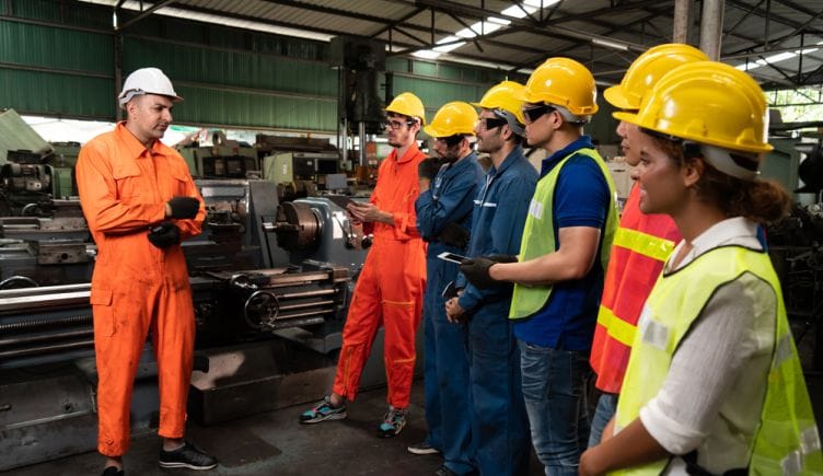 Several employees in hard hats and bright vests stand in an industrial setting listening to a trainer instruct them.