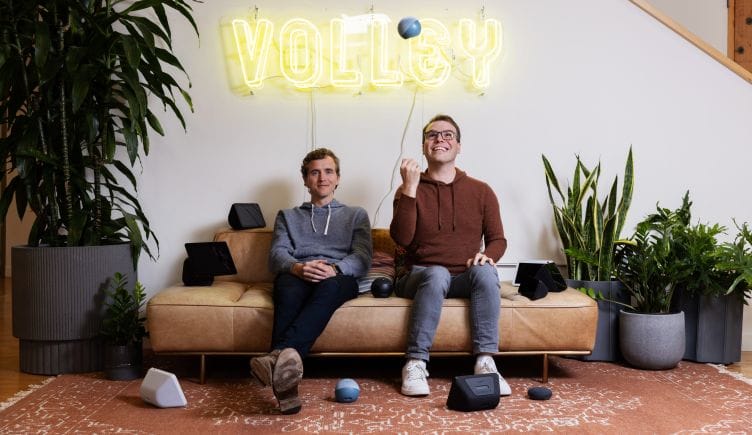 Two Volley teammates sit on a couch, one throwing a small ball in the air and smiling, with the word “Volley” in a neon sign on the wall behind them.