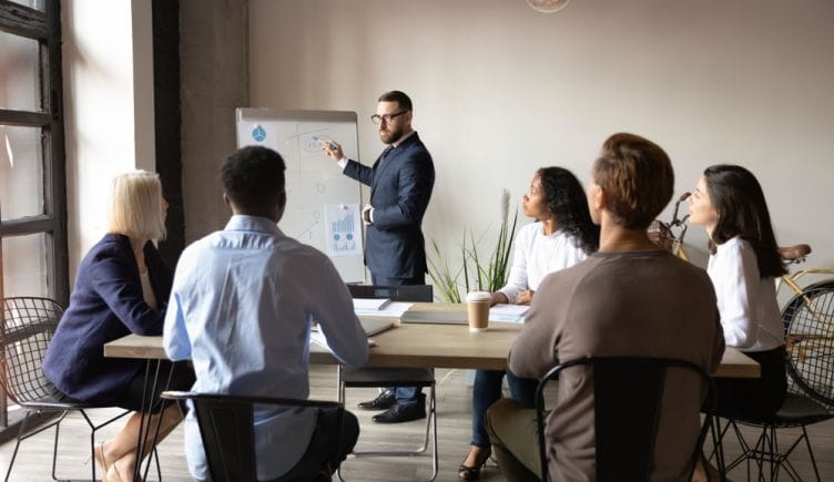 An instructor stands at the front of the room gesturing to a white board as he leads a training program for a group of professionals seated around a conference table.