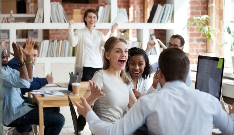 Several coworkers in a room together smile, clap and throw their hands in the air to represent employee engagement.
