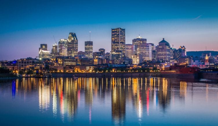 A view of the downtown Montreal skyline with several tall buildings lit up next to the water in early evening.