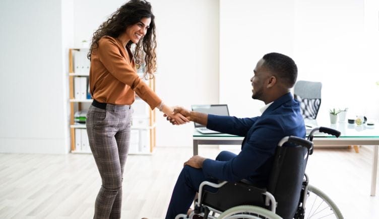 A businesswoman stands shaking hands with a businessman in a wheelchair.
