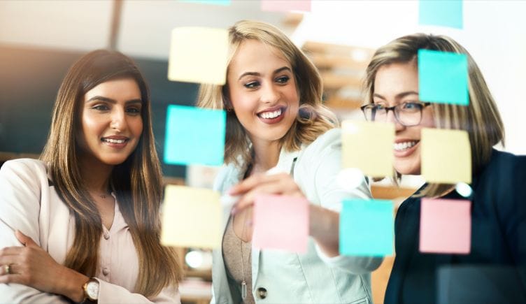 Three women coworkers in a planning session, smiling and looking at a clear board with colorful sticky notes