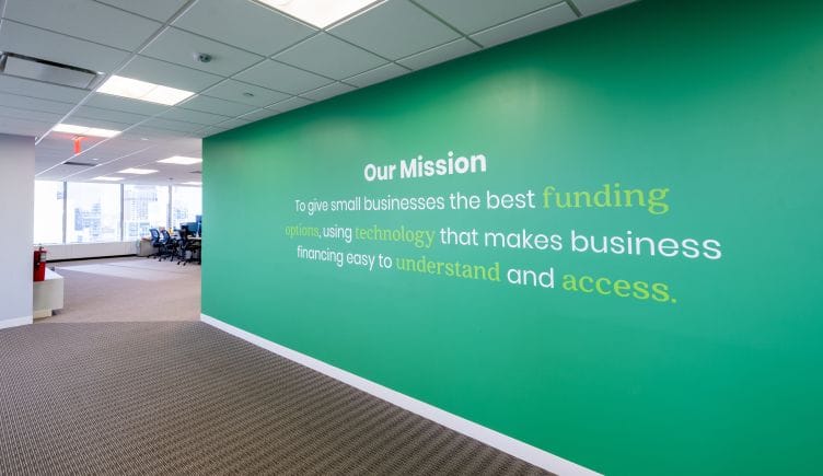 A green wall is printed with the text of Biz2Credit’s mission statement: “To give small businesses the best funding options, using technology that makes business financing easy to understand and access.” 