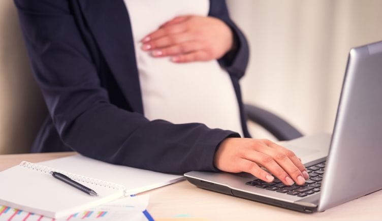 A close up of a pregnant person holding one hand over their stomach and using the other to type on a keyboard.