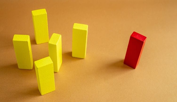 A red wooden block stands apart from a group of yellow wooden blocks, illustrating the marginalization that microaggressions cause in the workplace.