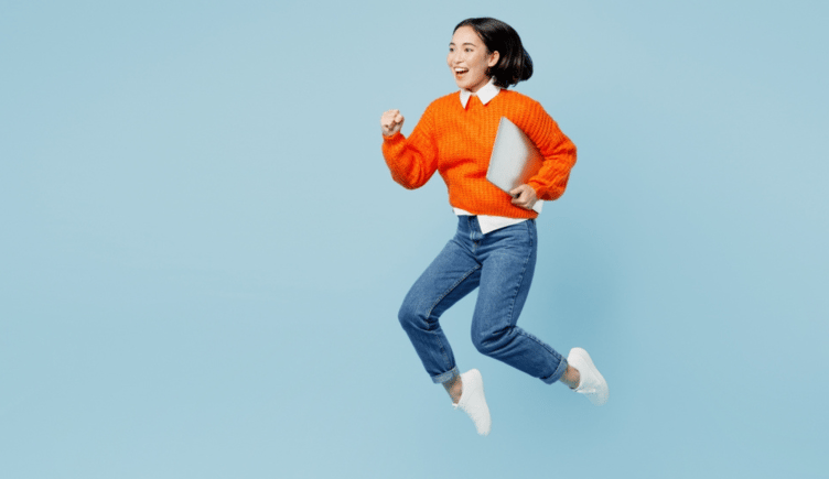 An excited young IT woman wears an orange sweater. She jumps while holding a laptop. 