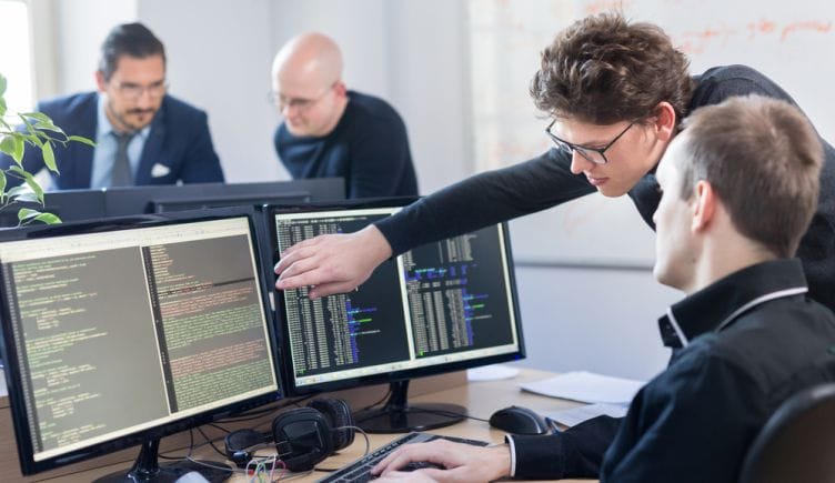 Two software professionals look over a pair of desktop computer monitors filled with lines of code.