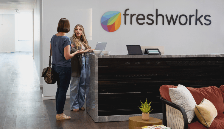 The front desk at Freshworks’ office, with logo wall and two employees standing and talking.