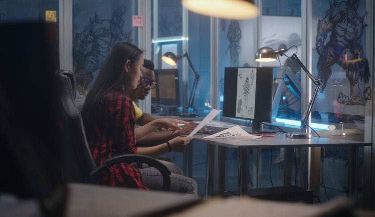 Two people sitting at a desk with a computer on it, discussing an image on both the computer and a piece of paper