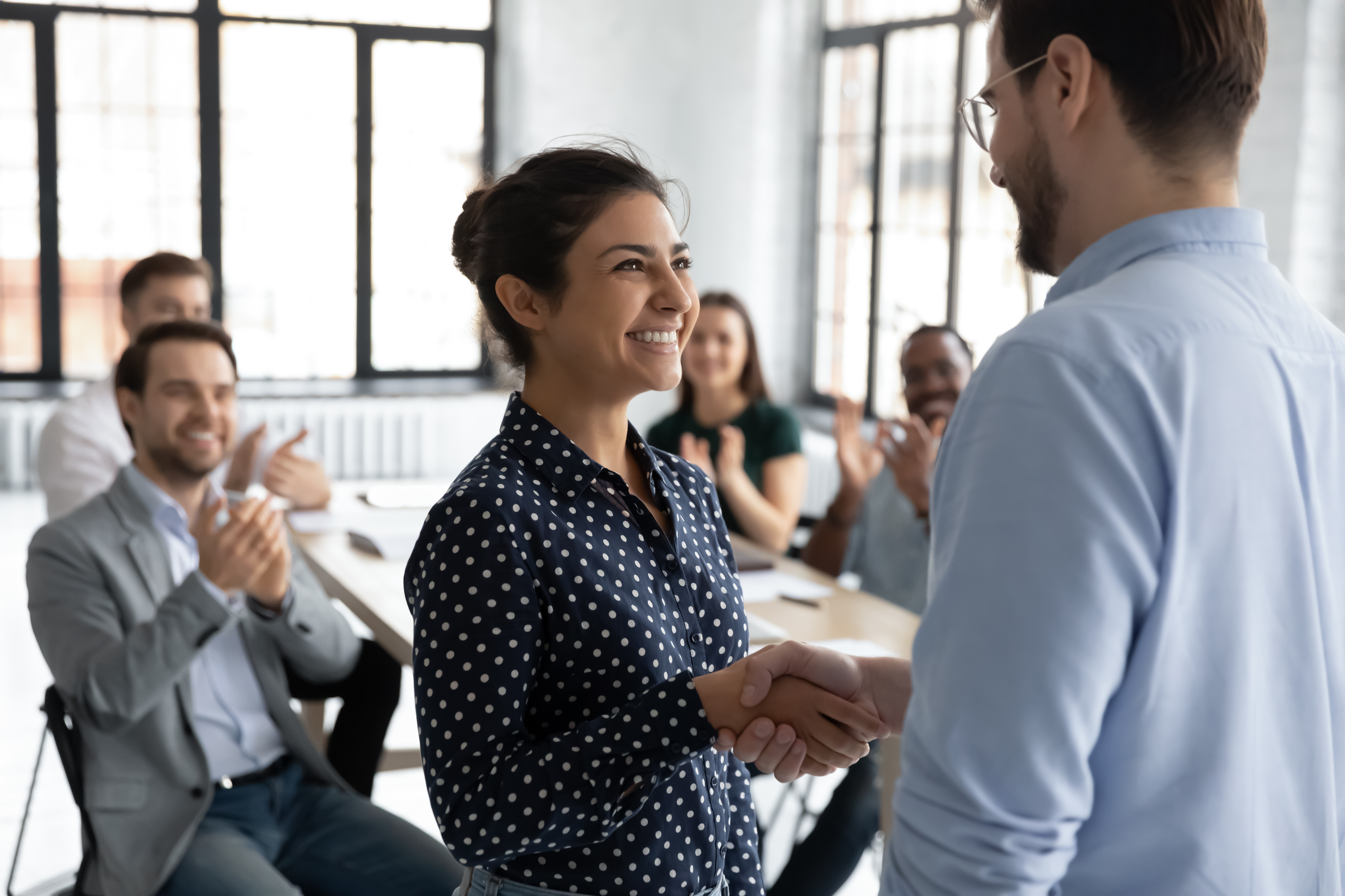 Young woman smiling and shaking the hand of a coworker with other coworkers seated behind them applaud her