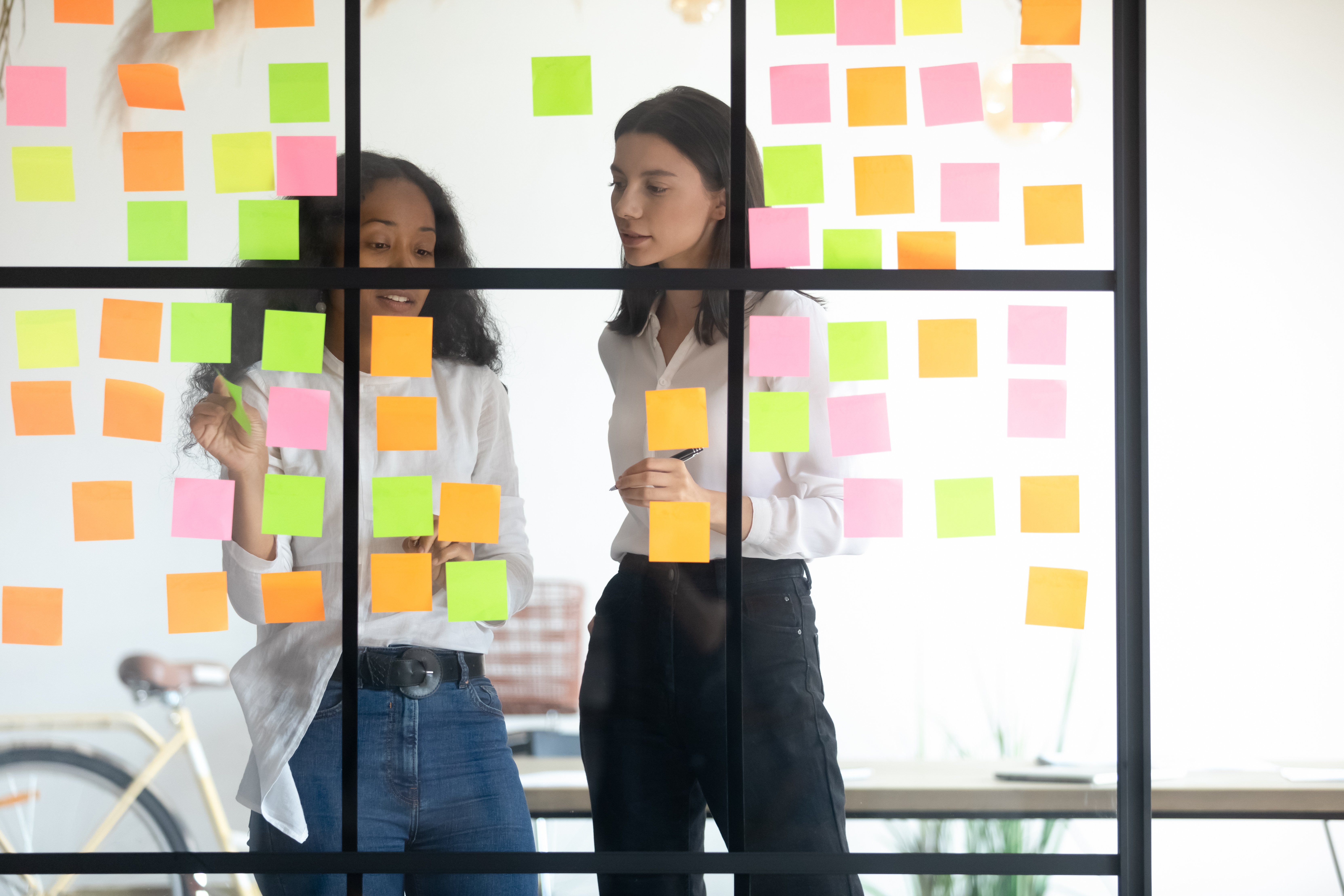 Image of coworkers discussing workflow laid out with sticky notes on a glass conference room wall.