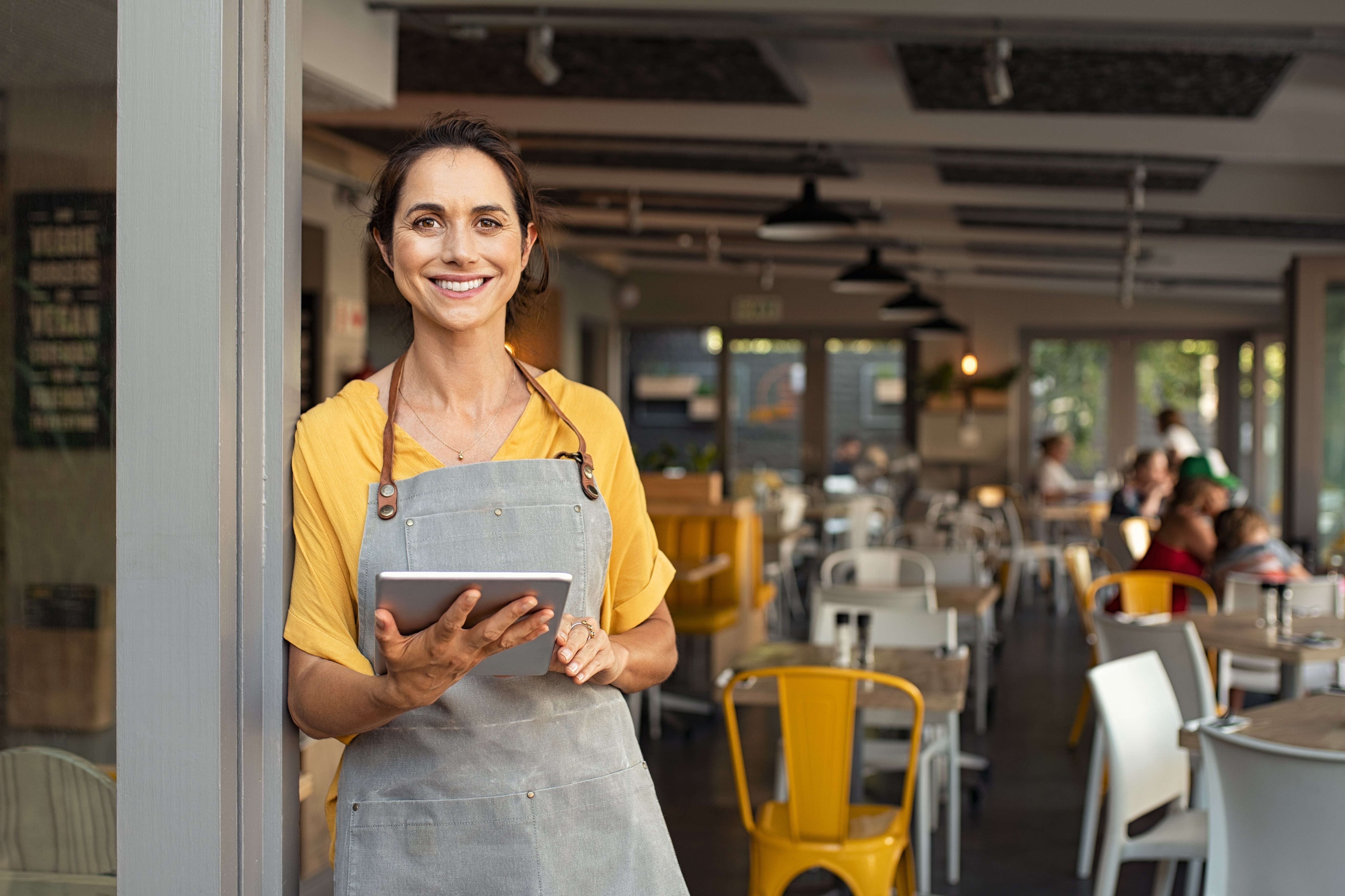 A woman in an apron leaning against the doorway of a restaurant, holding a tablet and smiling.
