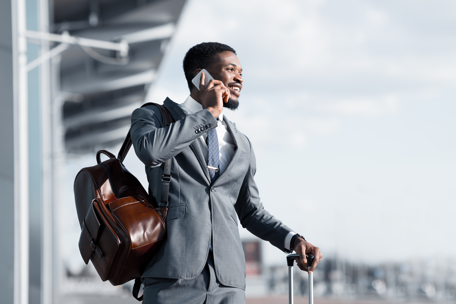 A Black man wearing a tailored suit talks on a phone at an airport while waiting for a taxi. He has a leather backpack slung over one shoul