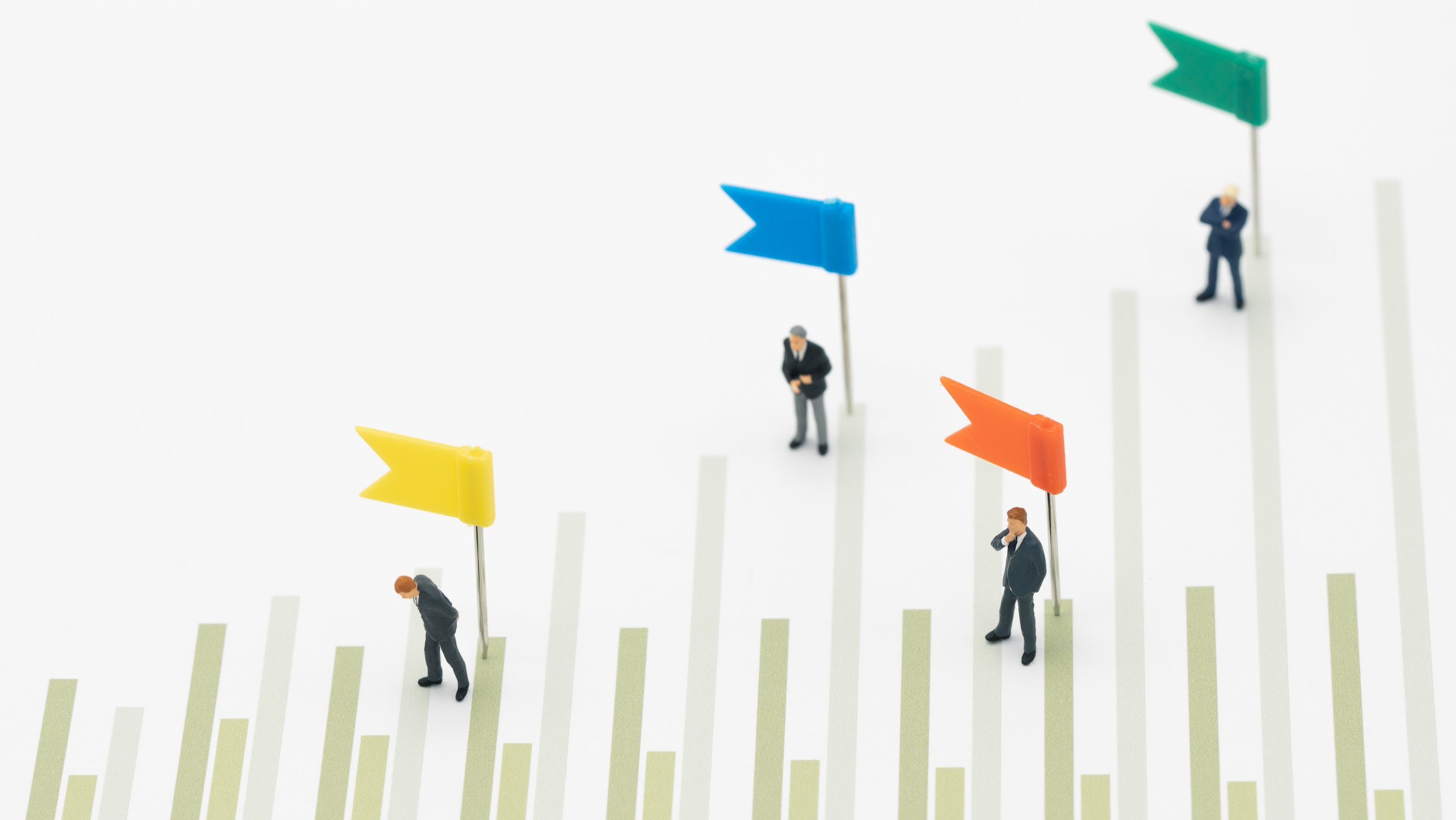 A photo of miniature people in suits standing on a piece of paper with a bar chart with colorful flags to note levels of achievement.