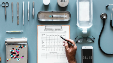 a doctor holds a pen over a clipboard. Various medical supplies are organized neatly on the table around the clipboard including a thermometer, an IV bag and a stethoscope.