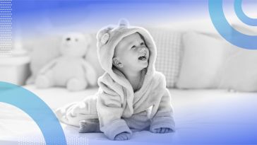 A baby laughs while sitting on a bed wearing a robe 
