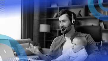 A bearded man wears a headset and holds a baby while talking into a computer. /remote-work/4-lessons-remote-working-parent