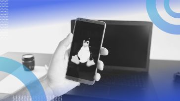 A hand holds a smartphone with the Linux logo in front of a laptop