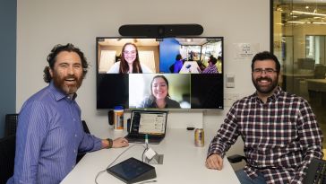 Bluecore's L&D team posing for a photo. Jason Arrigo and Christopher Carmena are on either side of a TV screen, sitting at a table. The TV screen shows a Zoom call of the two of them, Emily Nassir and one other employee.