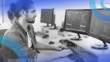 Ember.js image of a white man with a trimmed beard and his hair pulled back in a bun. He types code on a dual-monitor system. He wears a chambray shirt.