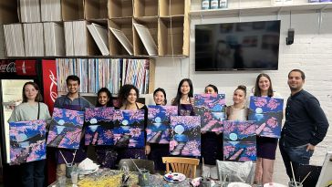 Some of Torch's employees posing for a photo, each holding their own painting of a body of water reflecting the moon and surrounded by cherry blossoms.