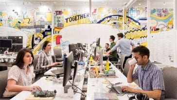 Photo of Zocdoc team members working at bench desk seating with wall full of graphic illustrations in background.