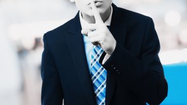 A person in a suit puts a finger to their lips as if to say, "quiet."