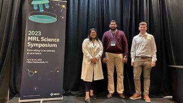 Three Merck team members stand next to banner for 2023 MRL Science Symposium 
