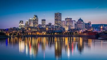 A view of the downtown Montreal skyline with several tall buildings lit up next to the water in early evening.