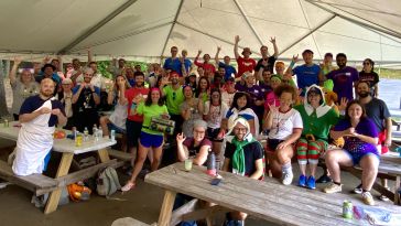  3Play team members are gathered around picnic tables under a big outdoor tent at a company event. They are wearing bright colors and have their hands up in the air.