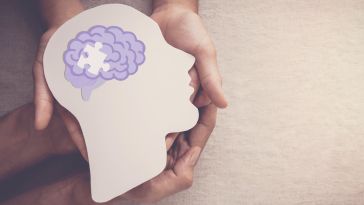 Photo of two sets of hands holding a paper cut-out of a head in profile that has a brain with a missing puzzle piece in the middle.