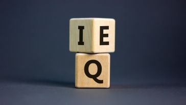 A wooden block with the letter "I" on one side and "E" on another side is stacked on top of a wooden block showing the letter "Q." The picture represents the two types of intelligence: emotional intelligence, which is measured as EQ, and cognitive intelligence, which is measured as IQ. 