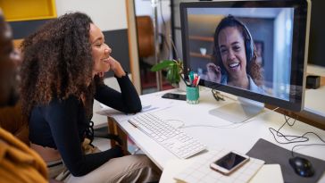 Two coworkers in an office speak with another colleague using video chat.