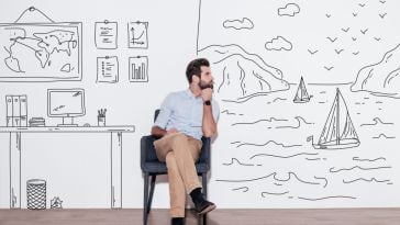 A man is seated in an office, with the walls behind him featuring drawings of a world map, charts and a seaside setting.